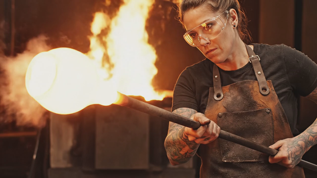 Macro cinematic shot inside a glassblowing workshop: molten glass glows and stretches as the artisan shapes a vase, furnace roar and crackle