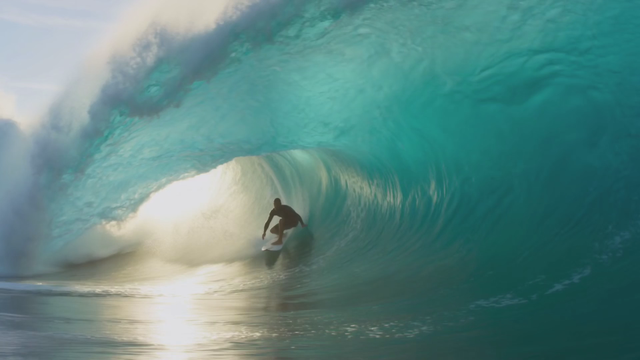 A surfer carves through the barrel of a turquoise wave in slow motion, spray glittering in backlight, camera tracking alongside inside the tube, roaring water fading to a muffled hush