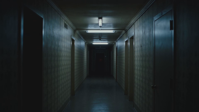 Handheld found-footage style shot of a long dark hallway, a single red balloon floats slowly toward the camera, flickering fluorescent light, low ominous hum