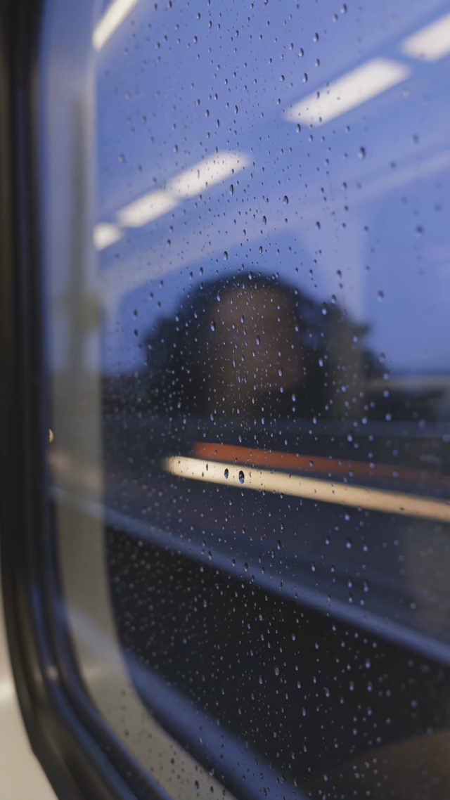 Close-up of raindrops racing down a train window at dusk, focus pulls to a passenger's reflection as city lights streak past, melancholic piano