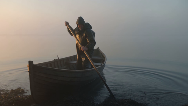 Three-shot sequence: a fisherman pushes his boat off a misty shore at dawn, then a wide aerial of the boat crossing glassy water, then a close-up of his hands pulling in a net heavy with fish, gulls and lapping water throughout