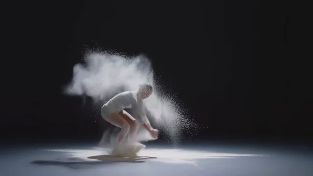 Một vũ công đương đại nhảy qua làn bột trắng trong một phòng tập tối, bột xoay quanh cơ thể cô trong chế độ quay chậm, một ánh đèn sân khấu đầy kịch tính, mọi hạt bột đều được chiếu sáng rõ nét
