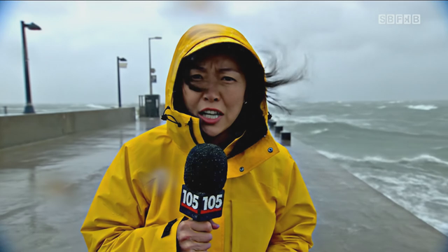 News-style stand-up: a reporter in a rain jacket speaks to camera on a windy pier, 'Conditions here are changing by the minute', waves crashing behind, handheld urgency