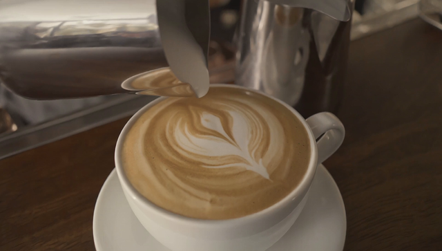 A barista slides a latte across the counter and says 'This one's on the house', cozy cafe ambience with espresso machine hiss and low chatter, warm tungsten light